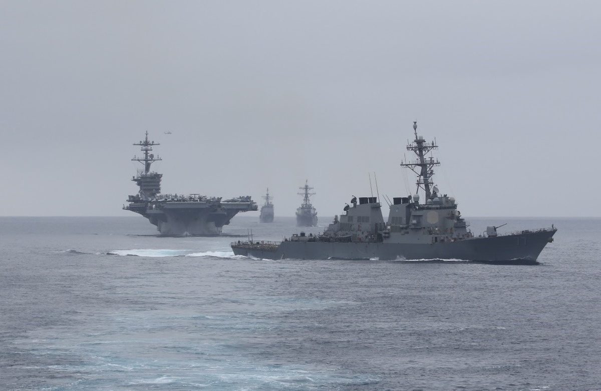 Ships of Carrier Strike Group 1 steaming in formation in the Pacific Ocean with aircraft overhead