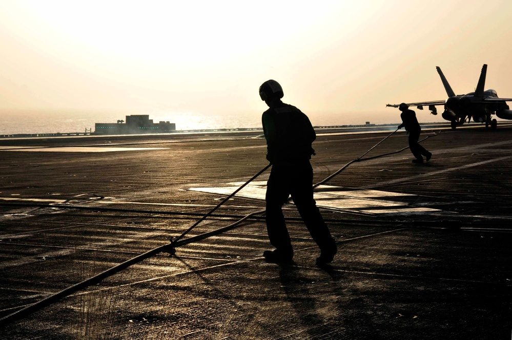 Sailors handling the cross-deck arresting wire on a carrier flight deck at sunset