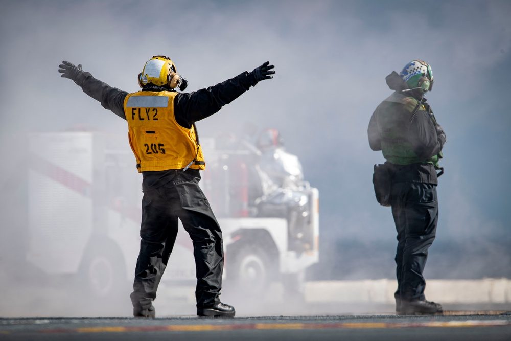 Yellow-shirted flight deck handler directing aircraft amid catapult steam with green-shirted crew alongside